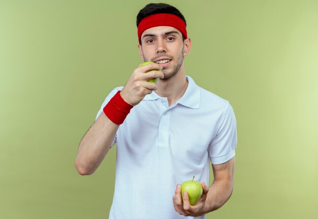 Jovem desportivo com uma bandana segurando maçãs verdes, mordendo uma em pé sobre um fundo verde