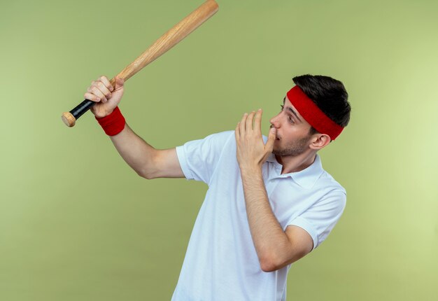 Jovem desportivo com uma bandana segurando um taco de beisebol olhando para ele espantado e surpreso em pé sobre um fundo verde