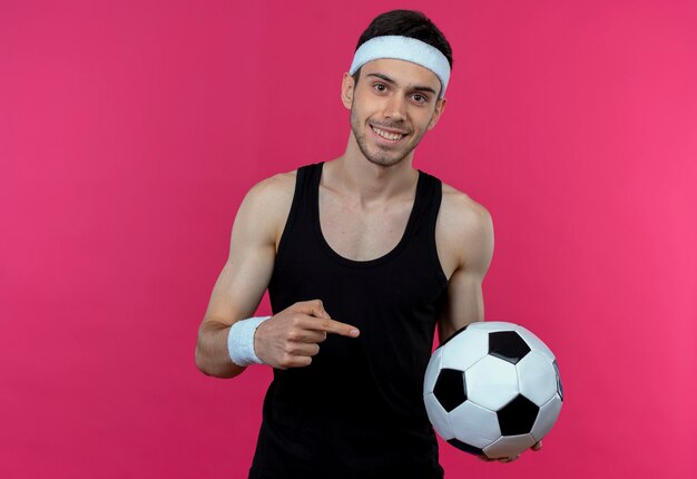 Jovem desportivo com uma bandana segurando uma bola de futebol, apontando com o dedo indicador para a bola, sorrindo feliz e positivo em pé sobre a parede rosa