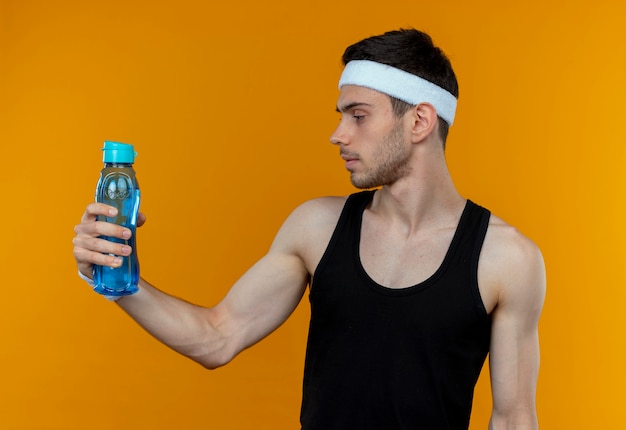 Jovem desportivo com uma bandana segurando uma garrafa de água e olhando para ela com uma cara séria em pé sobre a parede laranja