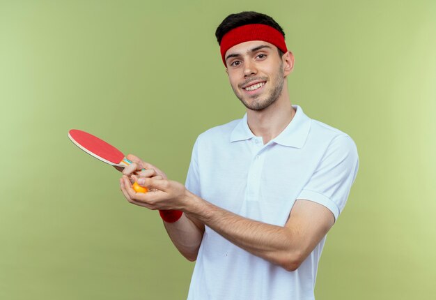 Jovem desportivo com uma bandana segurando uma raquete e uma bola de tênis de mesa, sorrindo em pé sobre a parede verde