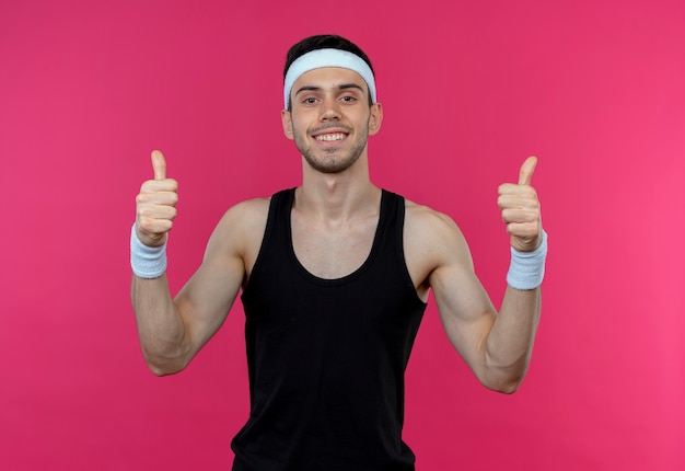 Jovem desportivo com uma bandana sorrindo, feliz e positivo, mostrando os polegares para cima sobre o rosa