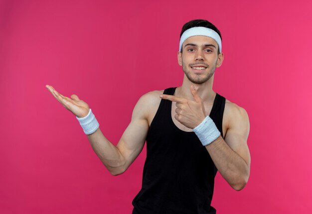 Jovem desportivo com uma faixa na cabeça, apresentando algo com o braço da mão apontando com o dedo para o lado, de pé sobre a parede rosa