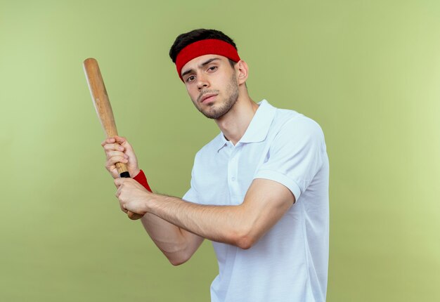 Jovem desportivo com uma faixa na cabeça, balançando o taco de beisebol, olhando para a câmera com uma cara séria em pé sobre um fundo verde