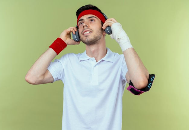 Jovem desportivo com uma faixa na cabeça com fones de ouvido e uma pulseira para smartphone feliz e positivo curtindo sua música sobre o verde