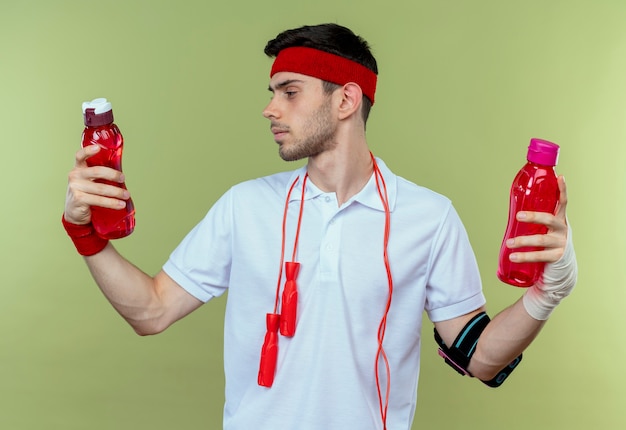 Foto grátis jovem desportivo com uma fita na cabeça e pular corda em volta do pescoço segurando duas garrafas de água, parecendo confuso tentando escolher o verde