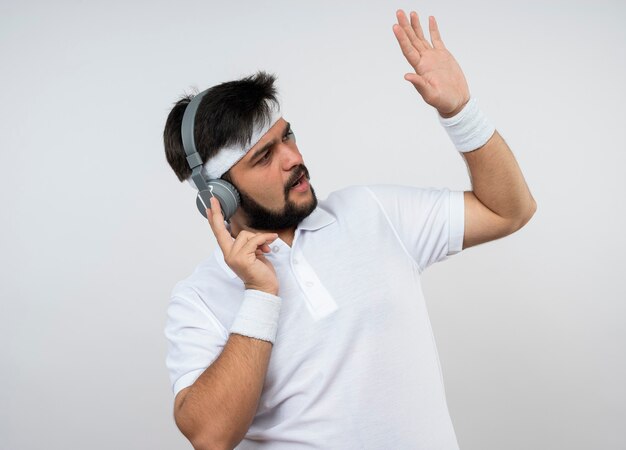 Jovem desportivo descontente, olhando para o lado usando bandana e pulseira com fones de ouvido, levantando a mão isolada na parede branca