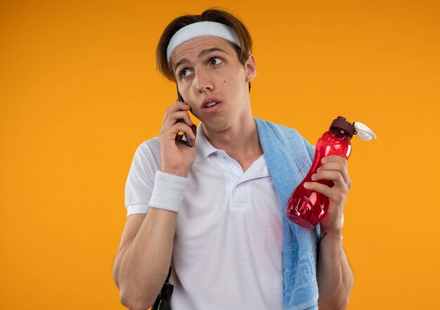 Jovem desportivo impressionado, olhando para o lado, usando bandana e pulseira com uma toalha no ombro falando ao telefone isolado na parede laranja