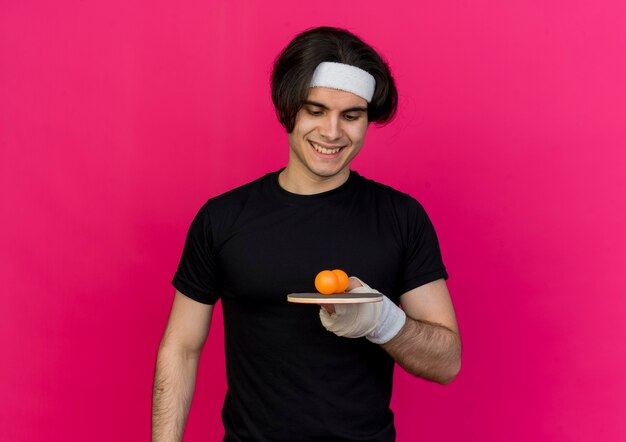 Jovem desportivo vestindo roupas esportivas e bandana segurando a raquete e a bola de tênis de mesa, sorrindo feliz e positivo