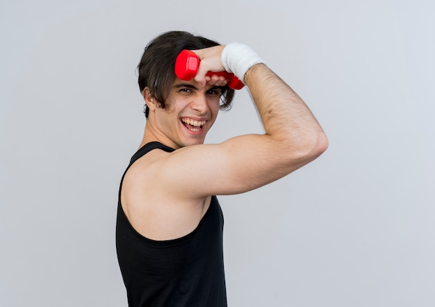 Foto grátis jovem desportivo vestindo roupas esportivas e bandana, segurando halteres, levantando a mão e sorrindo alegremente em pé sobre a parede verde