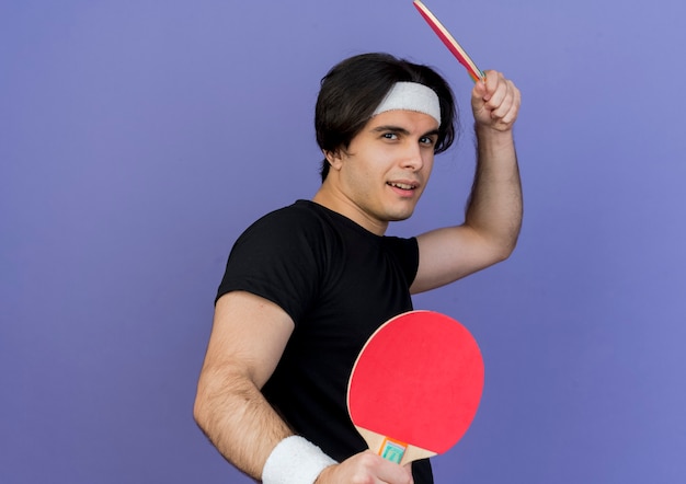 Jovem desportivo vestindo roupas esportivas e bandana segurando raquetes de tênis de mesa com cara séria