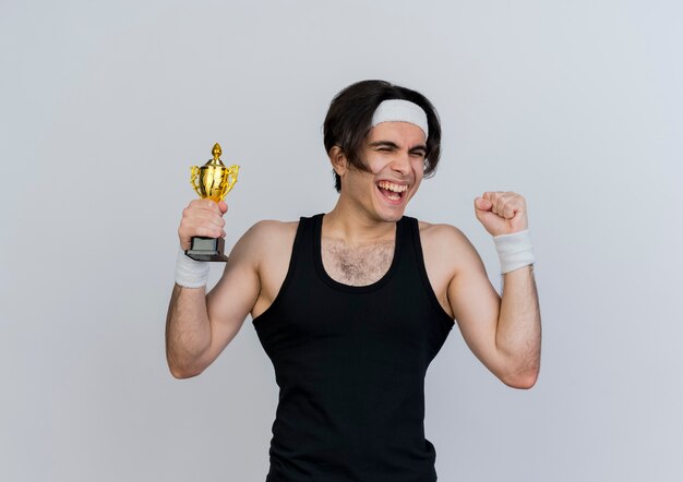 Jovem desportivo vestindo roupas esportivas e bandana segurando um troféu, feliz e animado, levantando o punho feliz e animado em pé sobre uma parede branca