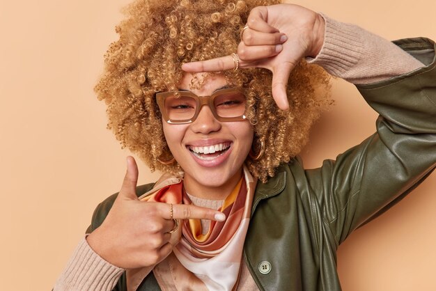 Jovem despreocupada positiva tem inspiração imagina como capturar tiro interessante mostra sorrisos de gesto de quadro com prazer usa óculos de roupas da moda isoladas sobre fundo marrom.