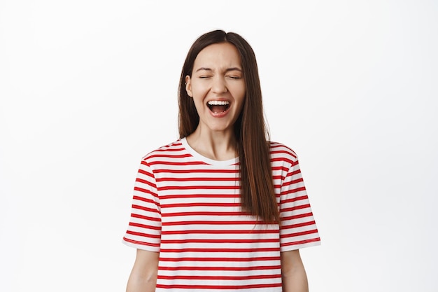 Jovem despreocupada, rindo e sorrindo, gritando de alegria e felicidade, de pé na camiseta listrada vermelha, roupas de verão, fundo branco.