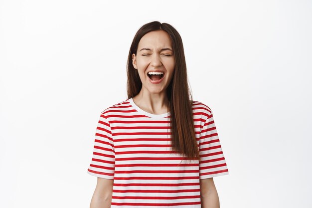 Jovem despreocupada, rindo e sorrindo, gritando de alegria e felicidade, de pé na camiseta listrada vermelha, roupas de verão, fundo branco.