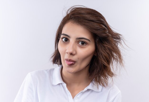 Jovem divertida e alegre com cabelo curto, usando uma camisa pólo branca, olhando para a câmera e mostrando a língua