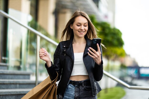 Jovem do lado de fora com sacolas de compras e usando o smartphone. Menina usa gadget digital, olhando na tela do smartphone.