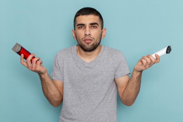 Jovem do sexo masculino com camiseta cinza segurando dois barbeadores elétricos diferentes no azul-gelo.