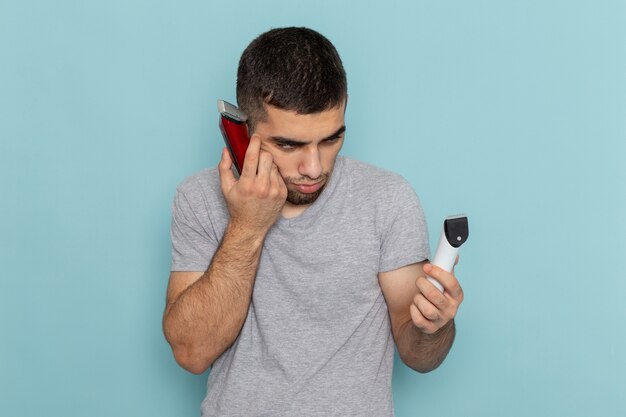 Jovem do sexo masculino com camiseta cinza segurando dois barbeadores elétricos diferentes, pensando em qual deles escolher