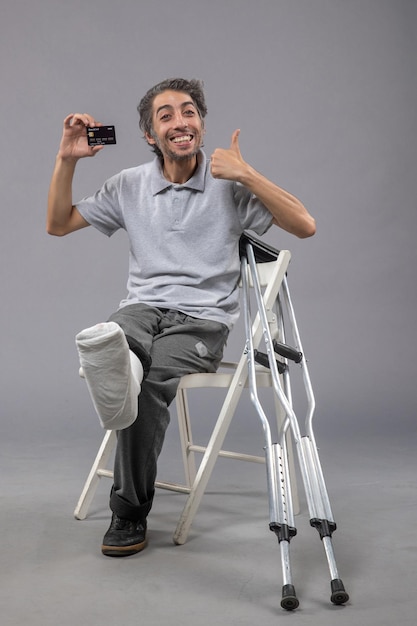 Foto grátis jovem do sexo masculino com o pé quebrado e curativo segurando o cartão do banco preto na parede cinza torção do sexo masculino com dor no pé acidente