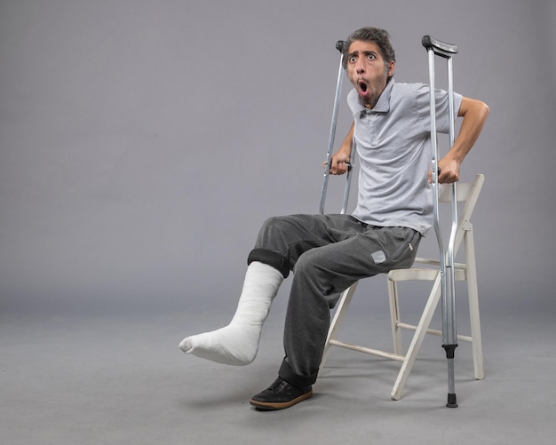 Jovem do sexo masculino com o pé quebrado tentando se levantar segurando muletas na parede cinza Pé quebrado dor torção acidente perna Vista frontal