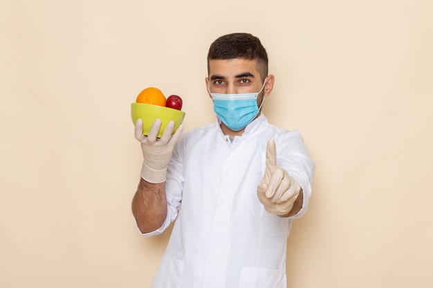 Jovem do sexo masculino de terno branco usando máscara e luvas segurando um prato com frutas em bege