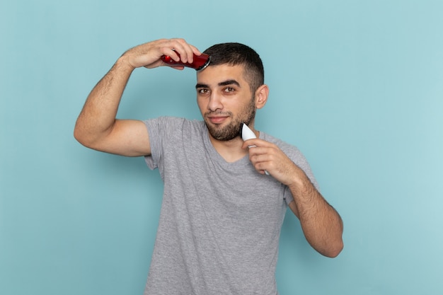 Jovem do sexo masculino em uma camiseta cinza segurando dois barbeadores elétricos diferentes, barbeando o rosto em azul-gelo, de frente