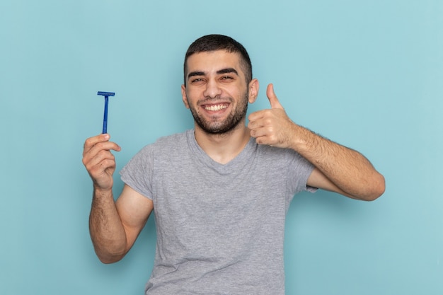 Jovem do sexo masculino em uma camiseta cinza segurando uma lâmina de barbear e sorrindo no azul de frente