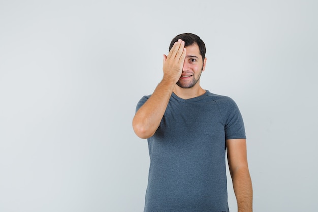 Jovem do sexo masculino segurando a mão em um olho em uma camiseta cinza e olhando positivo