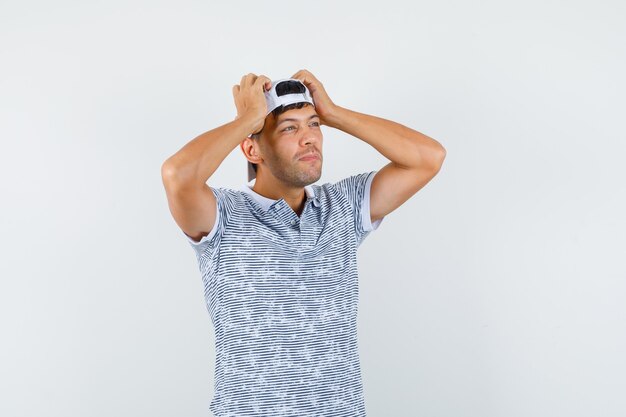 Jovem do sexo masculino segurando as mãos na cabeça com uma camiseta e boné e olhando pensativo