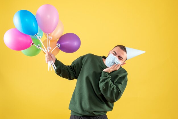 Jovem do sexo masculino segurando balões coloridos em um fundo amarelo de frente