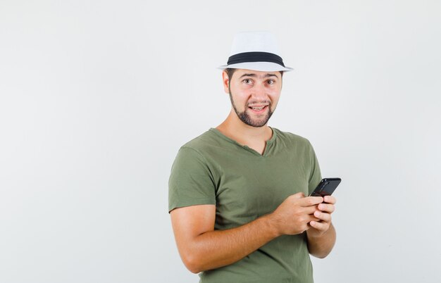 Jovem do sexo masculino segurando um celular com uma camiseta verde e um chapéu, parecendo feliz