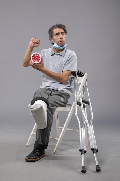 Jovem do sexo masculino sentado com atadura amarrada devido a um pé quebrado e segurando o relógio na parede cinza com dor no pé de torção humana