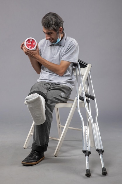 Jovem do sexo masculino sentado com atadura amarrada devido a um pé quebrado e segurando o relógio na parede cinza torção acidente na perna dor no pé masculino