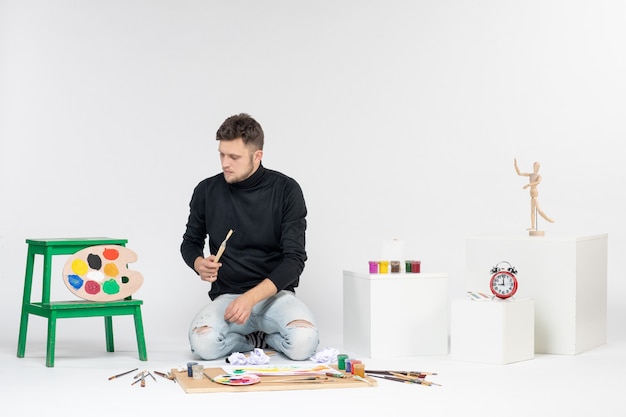 Jovem do sexo masculino sentado em frente a tintas segurando borlas para desenhar na parede branca pinturas de arte desenhar artista cor pintura