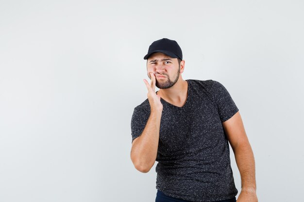 Jovem do sexo masculino sofrendo de dor de dente usando camiseta e boné, jeans e parecendo doente