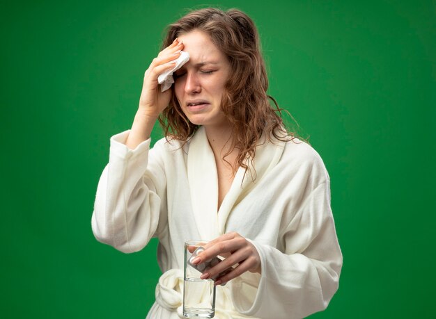 Jovem doente a chorar com os olhos fechados, usando um manto branco, segurando um copo de água com comprimidos e enxugando a testa com um guardanapo isolado no verde