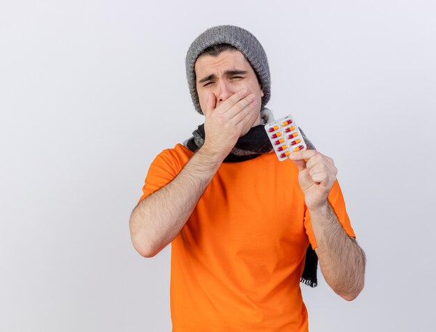 Jovem doente bocejando usando chapéu de inverno com lenço segurando comprimidos, boca coberta com a mão isolada no branco