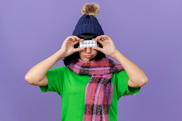 Jovem doente caucasiana com chapéu de inverno e lenço segurando um pacote de comprimidos na frente dos olhos isolados no fundo roxo