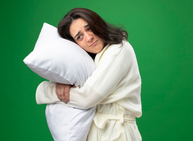 Foto grátis jovem doente caucasiana fraca vestindo manto em pé na vista de perfil, abraçando o travesseiro, olhando diretamente isolado sobre um fundo verde com espaço de cópia