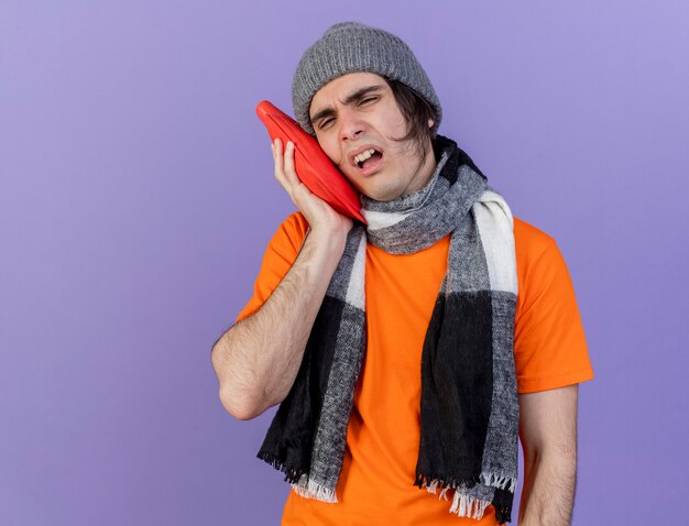 jovem doente com chapéu de inverno e lenço colocando uma bolsa de água quente na bochecha isolada no roxo