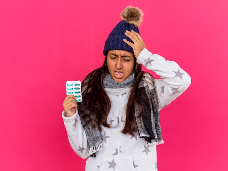 Foto grátis jovem doente com chapéu de inverno e lenço segurando comprimidos e colocando a mão na cabeça dolorida isolada no rosa