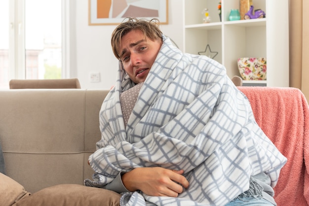 Jovem doente com dor e lenço em volta do pescoço envolto em um travesseiro xadrez, sentado no sofá da sala de estar