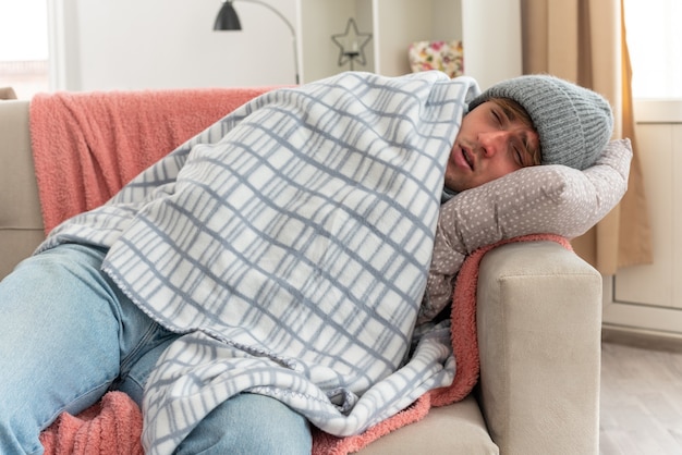 Foto grátis jovem doente com dor no pescoço e chapéu de inverno embrulhado em xadrez, deitado no sofá da sala de estar