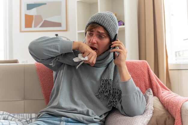 jovem doente com lenço no pescoço, chapéu de inverno, segurando a mão perto da boca e falando ao telefone, sentado no sofá da sala