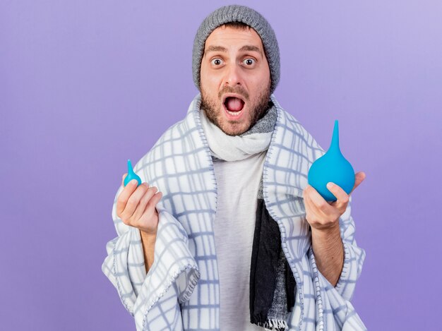 Jovem doente com medo usando chapéu de inverno com lenço segurando enemas isolados no fundo roxo