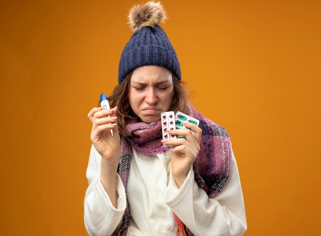 Foto grátis jovem doente com os olhos fechados, vestindo uma túnica branca e um chapéu de inverno com um lenço segurando um termômetro com comprimidos isolados na parede laranja