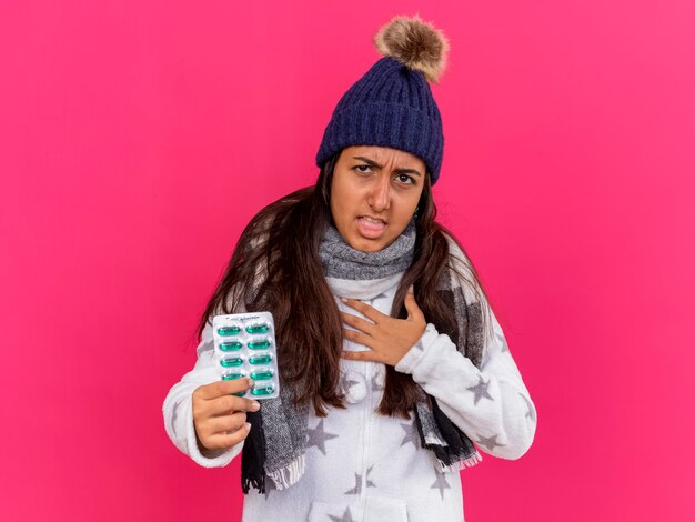 Jovem doente com tosse com chapéu de inverno e lenço segurando comprimidos colocando a mão no coração isolada em rosa