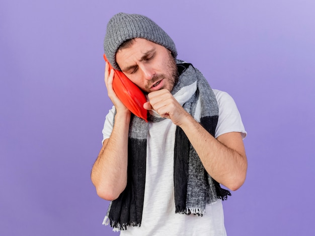 Jovem doente com tosse com chapéu de inverno e lenço segurando uma garrafa de água quente na bochecha isolada no roxo