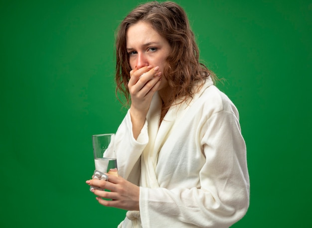 Foto grátis jovem doente com tosse, vestindo um manto branco, segurando um copo de água e colocando a mão na boca, isolado no verde